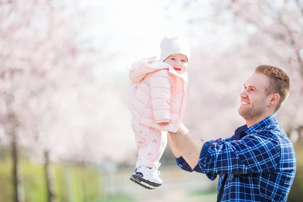 幼い父親は小さな可愛い娘を腕に抱えている 花の庭の父と娘 さくら — ストック写真