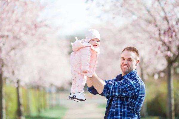 幼い父親は小さな可愛い娘を腕に抱えている 花の庭の父と娘 さくら — ストック写真