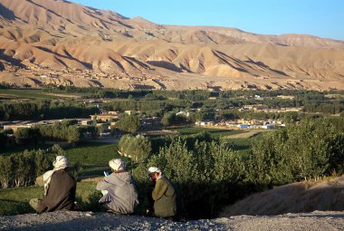 Orta Afganistan 'da Bamyan (Bamiyan). Üç Afgan adam bir tepede oturur ve ağaçlar ve uzak dağlarla Bamyan (Bamiyan) Vadisi 'nin üzerindeki manzaraya bakar. Bamyan (Bamiyan), Afganistan.