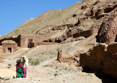 Orta Afganistan 'da Bamyan (Bamiyan). Bir kadın ve kız eve yürürken kafalarında mal taşırlar. Bu yerel evler Bamyan (Bamiyan) Buda 'larına yakındır. Afganistan 'da yerel hayat.