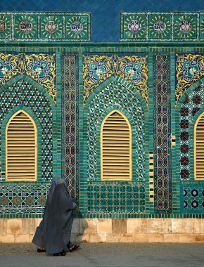 Afganistan 'ın Balkh ilindeki Mazar-i-Şerif' teki Mavi Cami. Mavi burka (burka) giyen iki kadın renkli fayanslar ve mozaiklerle süslenmiş caminin duvarının önünden geçiyor. Kuzey Afganistan.