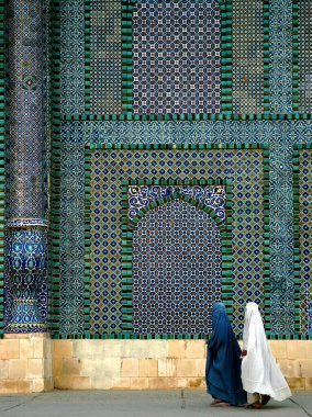 Afganistan 'ın Balkh ilindeki Mazar-i-Şerif' teki Mavi Cami. Burka giyen iki kadın renkli fayanslar ve mozaiklerle süslenmiş caminin duvarının önünden geçiyor. Kuzey Afganistan.