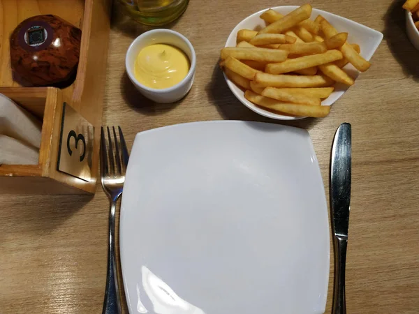 Çatal ve bıçaklı beyaz tabak. Ahşap masada soslu kızarmış patates..