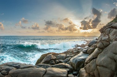 Gün batımında dalgalar kayalıklarda kırılır. Güneşin ışınları bulutları delip ıslak taşları aydınlatıyor. Sri lanka.