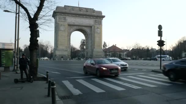 Bukarest / Románia - 03.05.2020: Forgalom a diadalív előtt (Arcul de Triumf), kora esténként.Egy elektromos robogóval várakozó férfi az utca túloldalán. — Stock videók