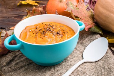 Sonbahar yapraklı ahşap masada, mavi bardakta, keten kumaşın üzerinde balkabağı resmi.