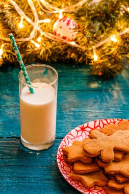 Noel kurabiyeli mavi masa fotoğrafı, bir bardak süt, yanan çelenkli ladin dalları.