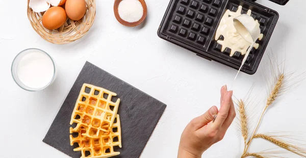 Photo on top of flour, eggs, waffle iron, spikelets, fresh roasted waffles, human hands on white table