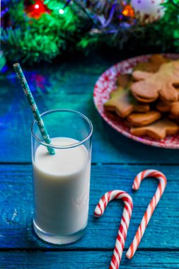 Üzerinde zencefilli bisküviler, bir bardak süt, karamel çubukları, yanan çelenkli ladin dalları olan mavi bir tablo.