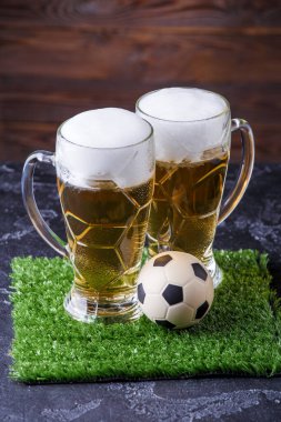 İki bardak bira, masada yeşil çimlerin üzerinde futbol topu.