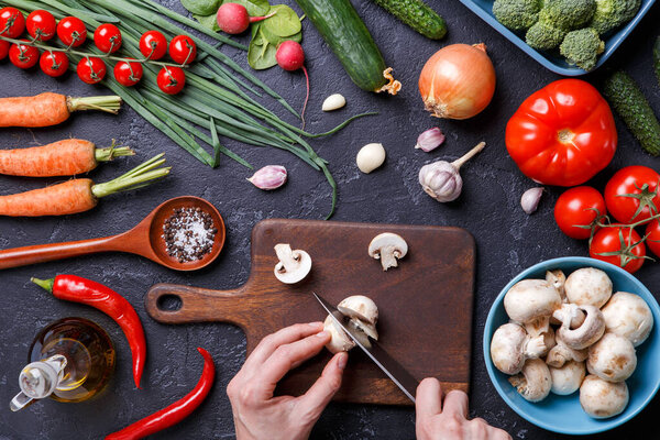 Picture on top of fresh vegetables, mushrooms, cutting board, oil, knife, hands of cook on table