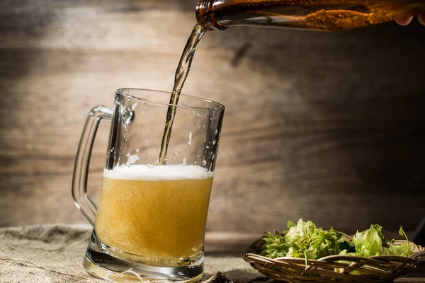 Foamy beer from bottle poured into mug standing near hops in basket