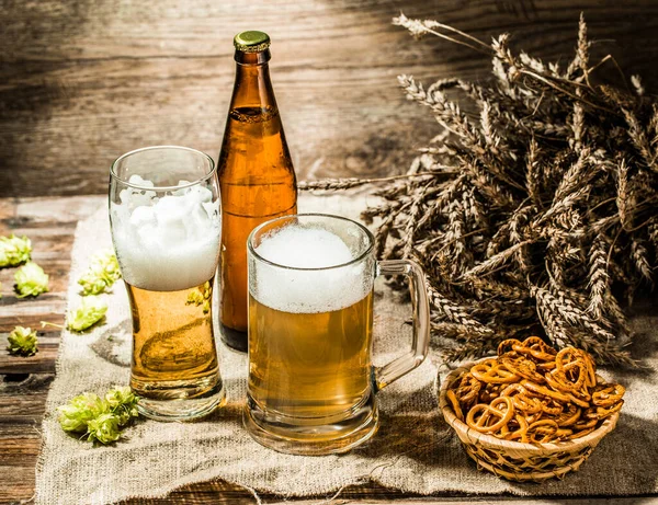 Mug, glasse, bottle of beer with foam on cloth with hop, pretzels and wheat