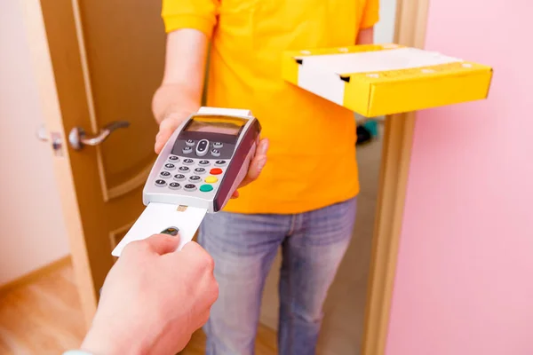 Photo of courier in yellow T-shirt with box with pizza and point of sale and buyer\'s hands with bank card on pink background