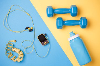 Fitness deneklerinin fotoğrafı. Oyuncu, dambıl, su şişesi, mavi ve sarı arka planda santimetre bant.