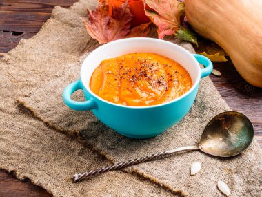 Portakallı balkabağı çorbasının üstünde fotograf, tencere, kaşık, sonbahar yaprakları ahşap masa kumaşı üzerinde.