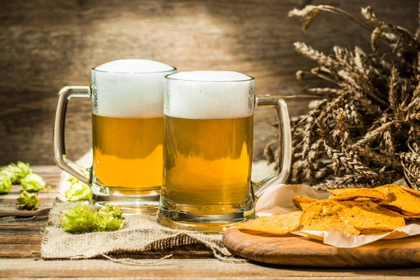 Two beer mugs with hops and chips on boards on linen cloth on wooden table
