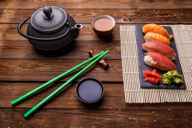 Suşi seti, soya soslu gri taş tahtada, gri çaydanlık ve ahşap zemin üzerinde bir fincan çay ile servis edilir.