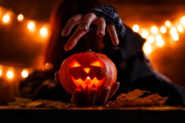 Arka planda yanan çelenklerle balkabağı tutan uzun saçlı bir cadı resmi