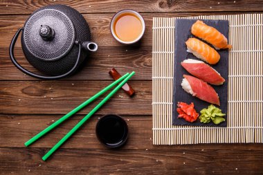 Suşi seti, soya soslu gri taş tahtada, gri çaydanlık ve ahşap zemin üzerinde bir fincan çay ile servis edilir.