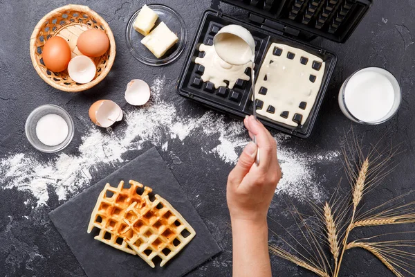 Photo on top of flour, eggs, waffle iron, spikelets, fresh roasted waffles, human hands on black table