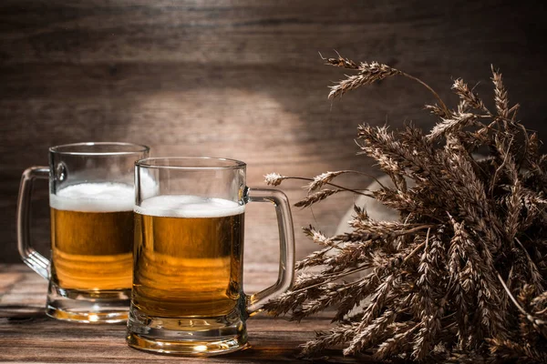 Two mugs of beer froth with wheat spikelets on empty wooden background