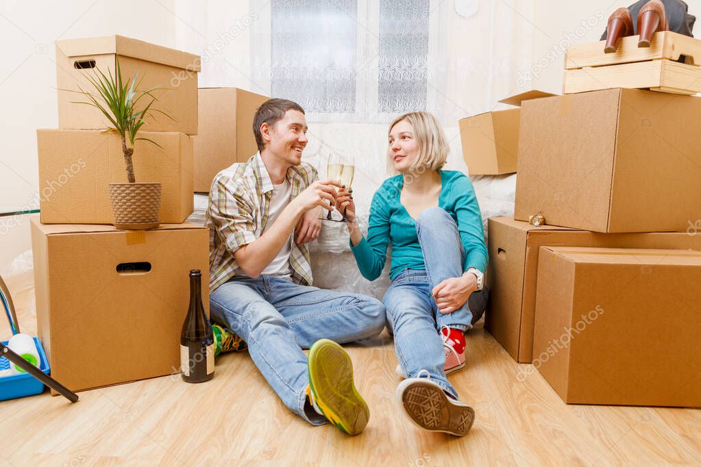 Photo of man with bottle of wine and woman sitting on sofa among cardboard boxes in new apartment