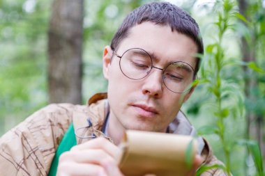 Koyu renk saçlı gözlüklü bir biyolog yaz öğleden sonra ağaçların ve bitkilerin arasında ormanda deftere yazıyor.
