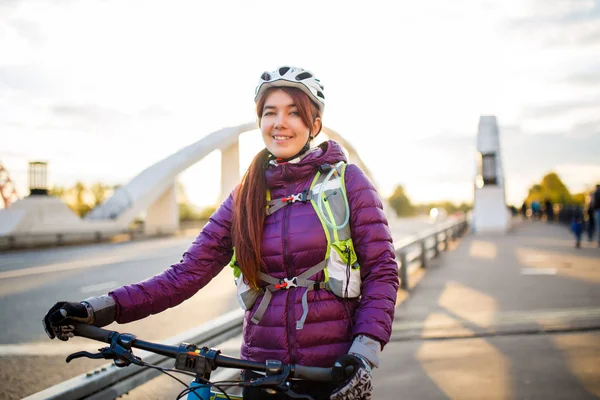 Photo of sportive woman in helmet on bicycle on bridge in city at autumn afternoon