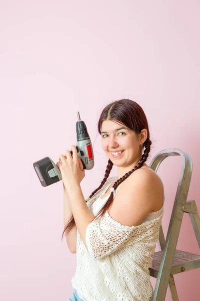 照片上是一个快乐的女人 在粉色墙壁的背景下 在梯子附近打钻 — 图库照片