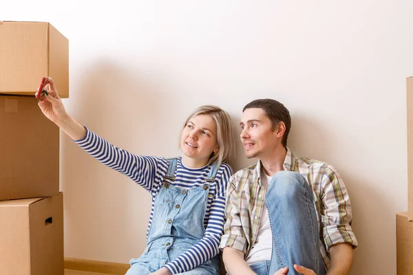 若い夫婦の写真新しいアパートの段ボール箱の間で床に座って自撮りをやって — ストック写真
