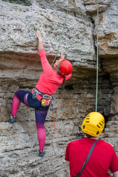 Photo from back of woman in helmet climbing on mountain and men with rope on summer day