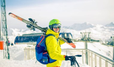 Omzunda kayaklarla gün boyunca kar kaplı fünikülerin arka planına bakan kasklı sporcu görüntüsü.