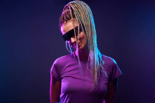 Donna con le trecce in occhiali da sole guardando la fotocamera in luce al neon — Foto Stock