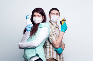 Tıbbi maske ve eldiven takmış bir kadın ve bir adam temas olmayan bir kızılötesi termometre ve antiseptik sabun tutuyorlar..