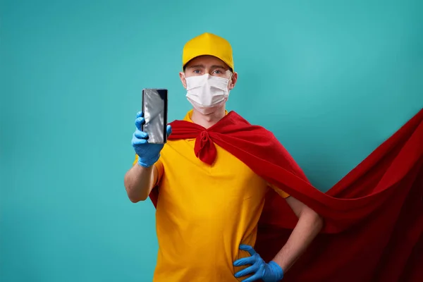 Courier dressed in superhero cloak and medical mask holds smartphone in his hand. — Stock Photo, Image