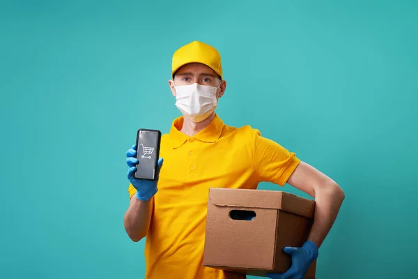 Kurýr s lékařskou maskou na tváři drží v ruce krabici a smartphone. — Stock fotografie
