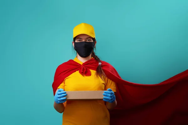 Курьер с коробкой одет в плащ супергероя и защищенную маску для лица . — стоковое фото