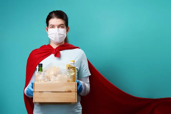 Dobrovolník na sobě chirurgické masky a superhrdina plášť drží jídlo box. — Stock fotografie