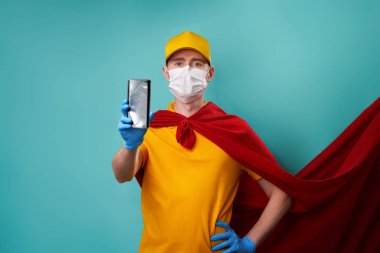 Süper kahraman pelerini ve sağlık maskesi takmış bir kurye akıllı telefonu elinde tutuyor..