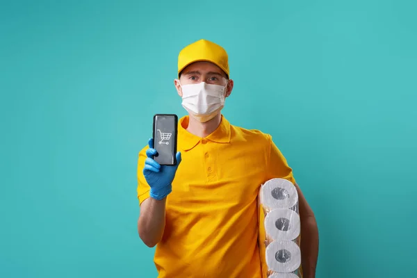 Courier está usando camiseta y gorra amarilla, máscara médica sostiene el teléfono inteligente en su mano y papel higiénico debajo del brazo . —  Fotos de Stock