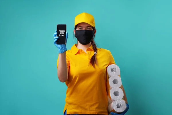 Courier está usando camiseta y gorra amarilla, máscara médica sostiene el teléfono inteligente en su mano y papel higiénico debajo del brazo . — Foto de Stock