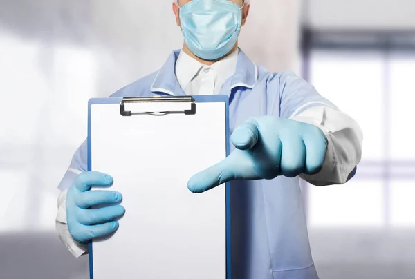 Photo of a doctor in suit, mask and rubber gloves holding a tablet for paper and pointing at camera.