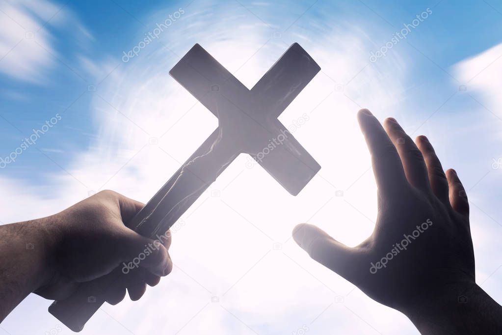 First person view photo of a male hands holding a cross on a bright light shine with blue skies background.
