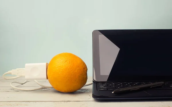 Orange fruit plugged in table laptop laying on a grey wooden table surface with green background.