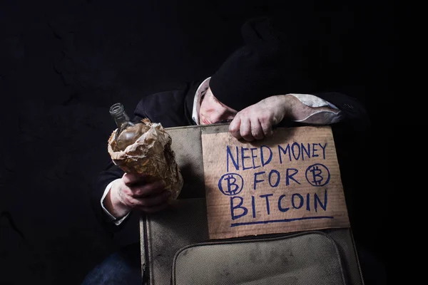 Photo of a drunk homeless businessman sleeping on travel suitcase with empty bottle and cardboard bitcoin sign.