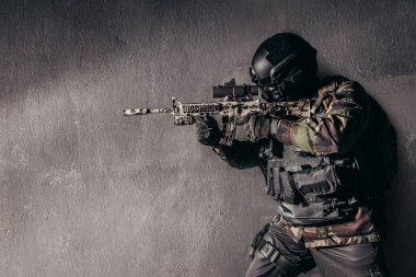 Kamuflaj üniformalı tam teçhizatlı bir askerin kirli beton arka planda duran ve nişan alan bir tüfeğin fotoğrafı..