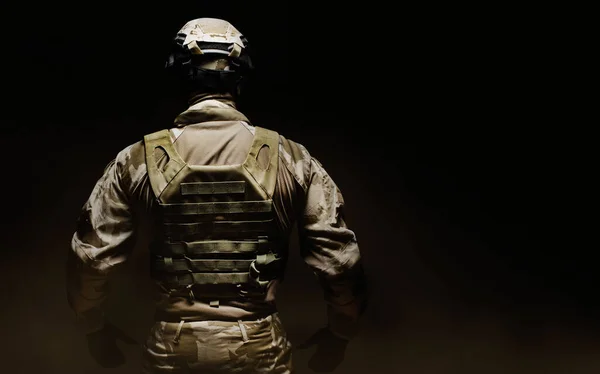 Photo of a fully equipped desert camouflage soldier in mask, helmet, armor and gloves on black foggy background back view.