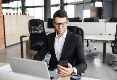 Bilgisayarda çalışan bir iş adamı. Ofiste dizüstü bilgisayar ve akıllı telefon kullanan genç bir adam. Yönetim, finans, iş kavramı