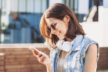 Kulaklıklı genç kız dışarıda akıllı telefon kullanıyor.